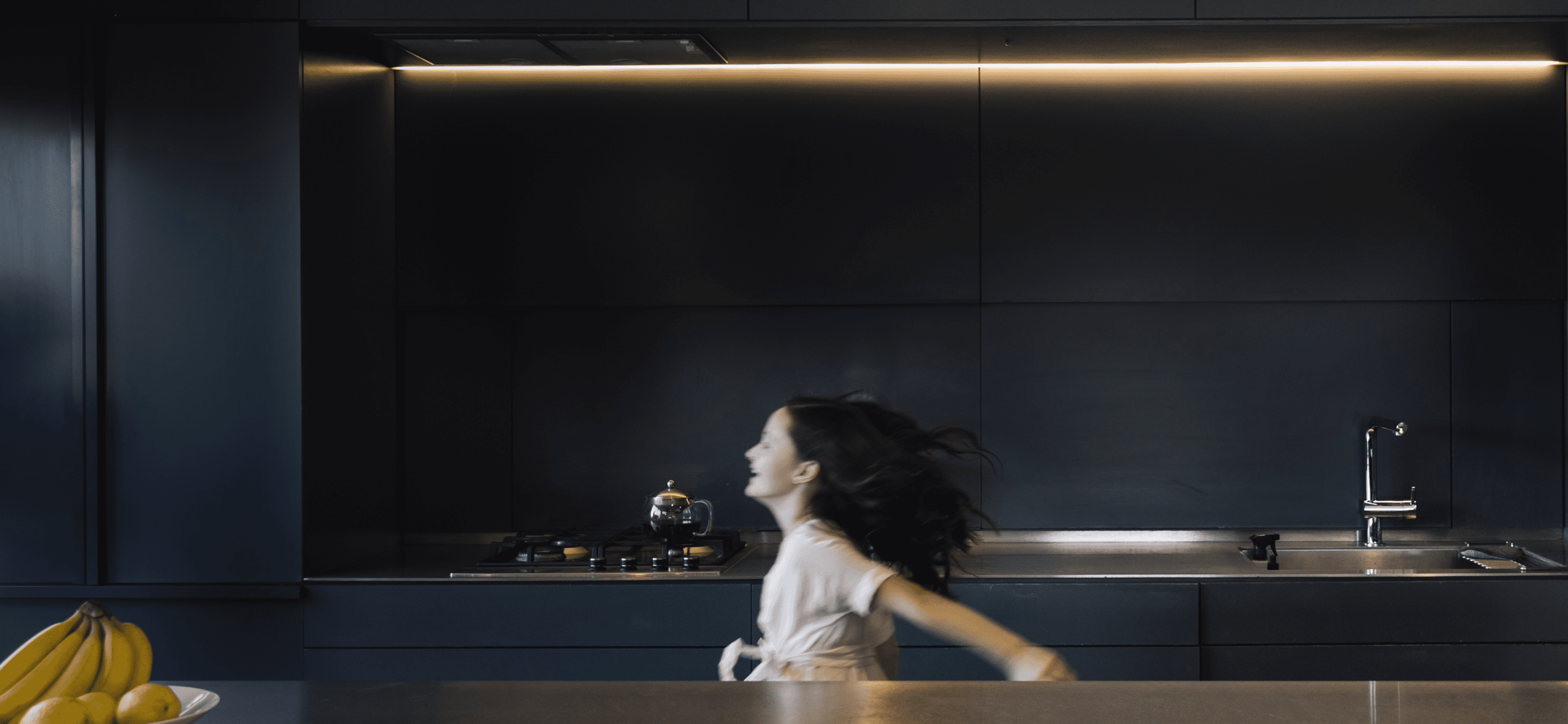 A blurred young girl in motion, adding a sense of liveliness and action to the sleek, modern kitchen with its dark cabinets and ambient lighting.