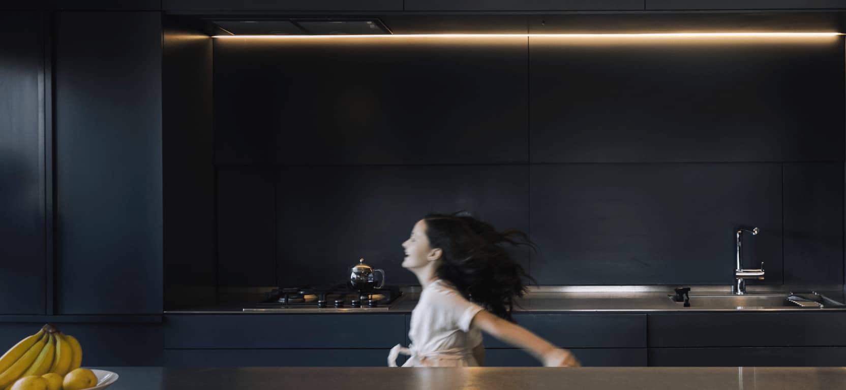 A young girl in a white shirt passing by a sleek modern kitchen with motion blur, indicating movement and life within a contemporary home setting.